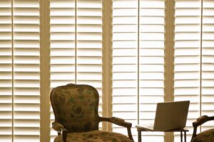 Brown armchair in front of plantation shutters | Featured Image for the PVC v Timber Shutters Blog by Shutters Blinds & Awnings.